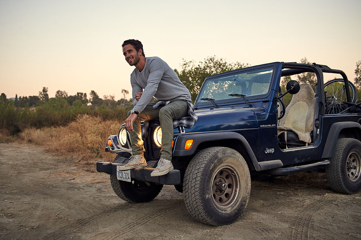 Man sitting on hood of blue jeep at sunset wearing PX Clothing long sleeve shirt and pans