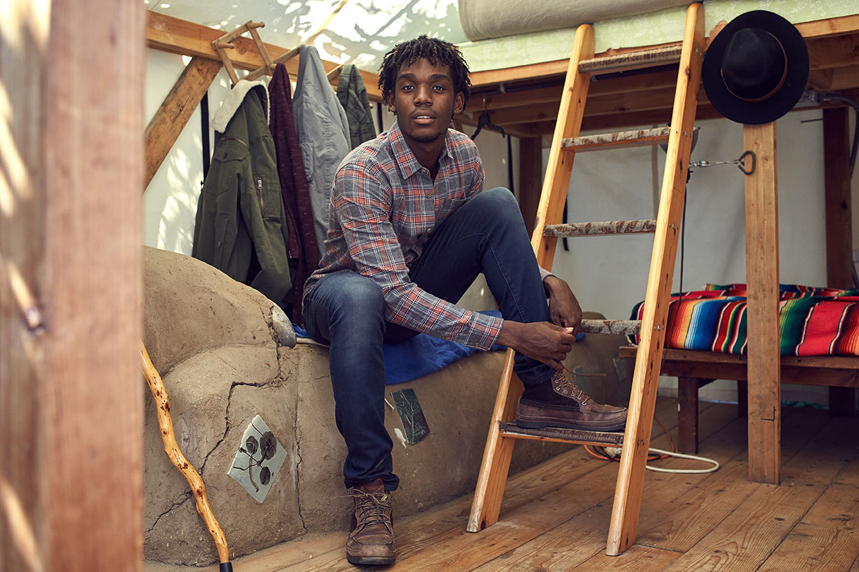 Man in purple plaid collared shirt, blue jeans, and boots from PX clothing sitting next to ladder